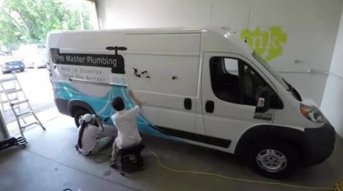 Two people working on a white van with 'Master Plumbing' branding in a garage.