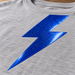 AI application illustration: glossy blue metal-flake-style lightning graphic on gray fabric; representative finish and color.
