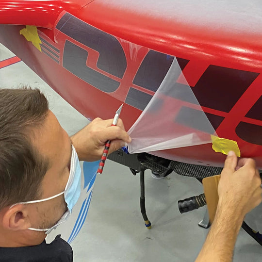 A man wearing a light blue face mask applies a transparent protective film over a red and black graphic decal on a vehicle body panel, using a pencil and tape to hold the film in place.