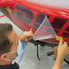 A man wearing a light blue face mask applies a transparent protective film over a red and black graphic decal on a vehicle body panel, using a pencil and tape to hold the film in place.