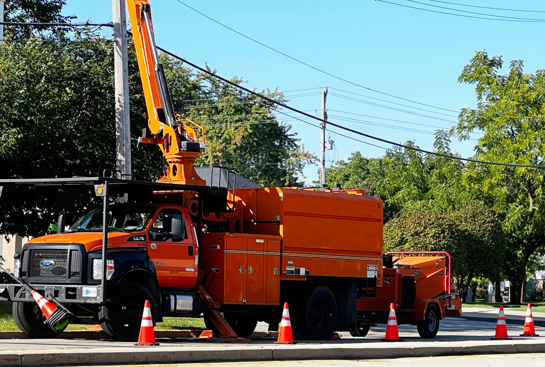 Orange utility truck with crane on a street with traffic cones and trees in the background