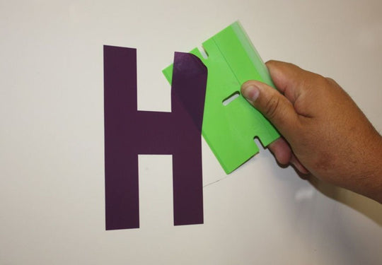 Hand holding a green letter 'H' cutout next to a purple letter 'H' on a white background