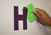 Hand holding a green letter 'H' cutout next to a purple letter 'H' on a white background