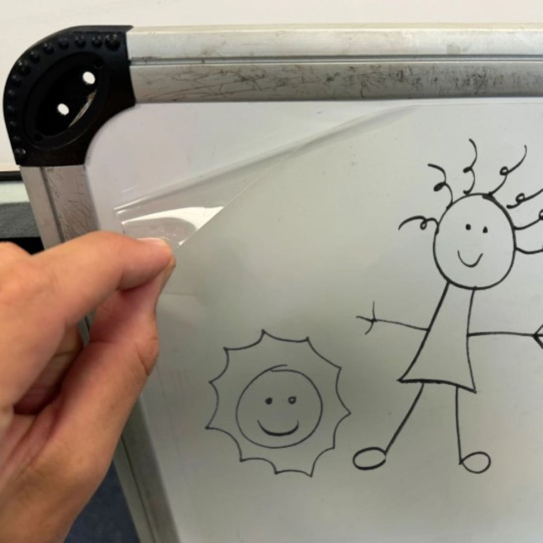 Hand holding a clear plastic sheet over a whiteboard with stick figure drawings of a girl and a sun.