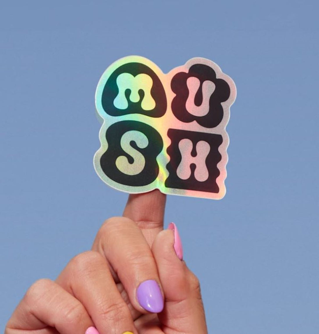 Holographic 'MUSH' sticker held by a hand against a blue background