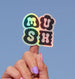 Holographic 'MUSH' sticker held by a hand against a blue background
