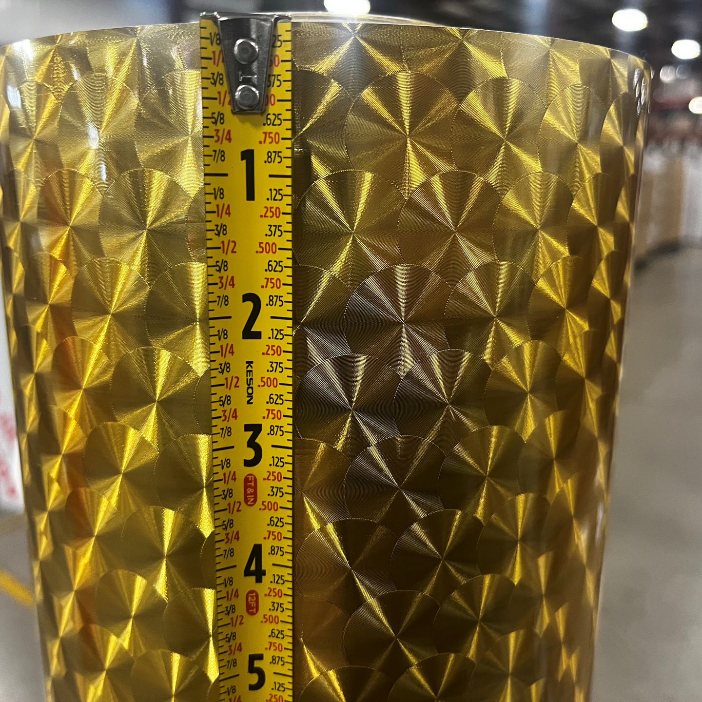 A close-up of a metallic gold textured bucket or planter with a 3D geometric starburst pattern, shown with a measuring tape vertically aligned on the front to indicate its height.