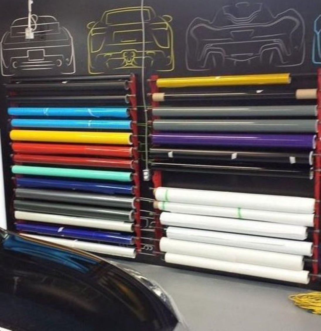 Rolls of vinyl wrap in various colors on a rack with car designs in the background.