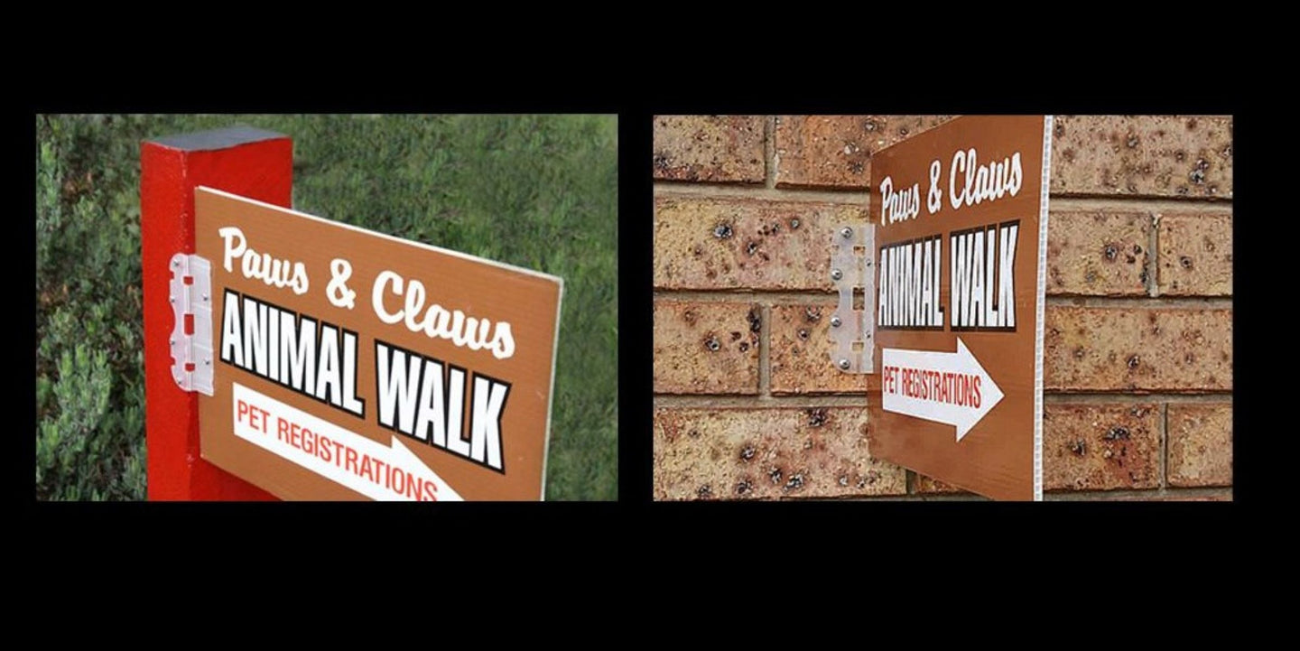 Two signs for 'Paws & Claws Animal Walk' on a grassy area and brick wall.