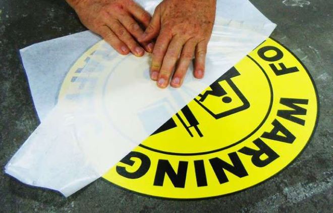 Yellow warning sign with black text and a circular design, held by hands on a dark surface.