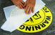 Yellow warning sign with black text and a circular design, held by hands on a dark surface.